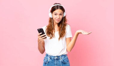 Genç ve güzel bir kadın, kafası karışmış, kulaklık ve akıllı telefonlardan şüphe duyuyor.