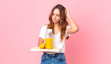 Genç, güzel bir kadın kafası karışmış ve kafası karışmış hissediyor. Kafasını kaşıyor ve elinde portakal suyuyla bir tepsi tutuyor.