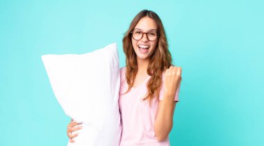 Genç ve güzel bir kadın şok geçiriyor, gülüyor ve başarıyı kutluyor pijama giyiyor ve yastık tutuyor.
