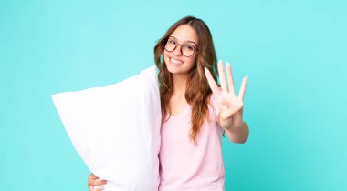 Genç, güzel bir kadın gülümsüyor ve arkadaş canlısı görünüyor. Dört numara pijama giyiyor ve yastık tutuyor.