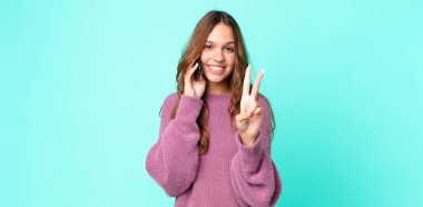 Genç, güzel bir kadın gülümsüyor ve arkadaş canlısı görünüyor. İki numarayı gösteriyor ve akıllı telefon kullanıyor.