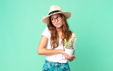 Genç, güzel bir kadın omuz silkiyor. Kafası karışık ve emin değil. Hasır şapkası var ve elinde kaktüs tutuyor.