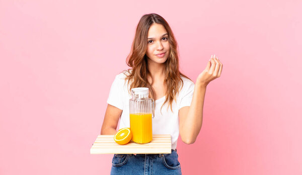 young pretty woman making capice or money gesture, telling you to pay and holding a tray with an orange juice