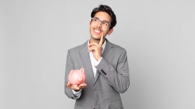 Genç İspanyol adam mutlu bir şekilde gülümsüyor ve hayal kuruyor ya da kuşkulanıyor ve kumbarasını tutuyor.