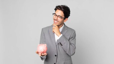 Genç İspanyol bir adam mutlu, kendinden emin bir ifadeyle gülümsüyor. Elleri çenesinde ve kumbarasında.