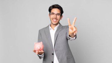 Genç İspanyol adam gülümsüyor ve mutlu görünüyor, zafer ya da barış işareti yapıyor ve kumbarasını tutuyor.
