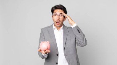 Genç İspanyol adam mutlu görünüyor, şaşırmış ve şaşırmış ve elinde bir kumbara tutuyor.