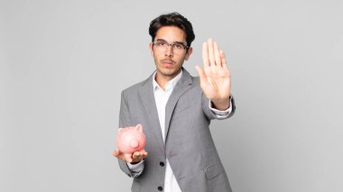 Genç İspanyol adam ciddi görünüyor. Avuç içi gösteriyor. Jest yapıyor ve elinde kumbara tutuyor.