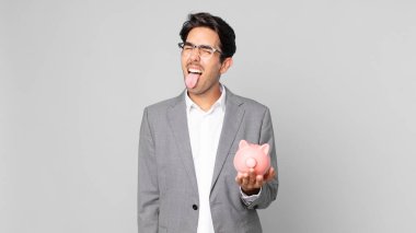 Neşeli ve asi tavırları olan genç İspanyol bir adam, şaka yapıyor, dilini çıkarıp domuz kumbarasını tutuyor.