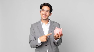 Genç İspanyol adam neşeyle gülümsüyor, mutlu hissediyor ve bir kenarı işaret ediyor ve kumbarayı tutuyor.