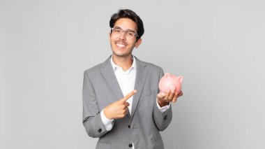 Genç İspanyol adam heyecanlı ve şaşırmış görünüyor. Bir tarafı işaret ediyor ve kumbarayı tutuyor.