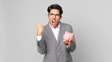 Genç İspanyol adam öfkeli bir ifadeyle bağırıyor ve elinde domuz kumbarası tutuyor.