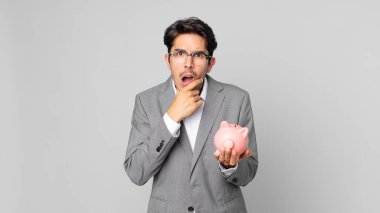 Ağzı ve gözleri açık, eli çenesinde ve elinde domuz kumbarası olan genç İspanyol bir adam.