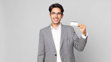 Genç İspanyol adam mutlu bir şekilde gülümsüyor, eli kalçasında, kendinden emin ve elinde bir kredi kartı var.