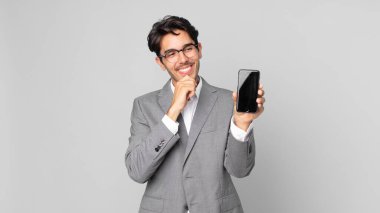 Genç İspanyol bir adam mutlu, kendinden emin bir ifadeyle gülümsüyor. Çenesi çenesinde ve elinde akıllı bir telefonla.