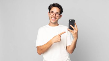 Genç İspanyol adam neşeyle gülümsüyor, mutlu hissediyor ve bir kenarı işaret ediyor ve elinde akıllı bir telefon tutuyor.
