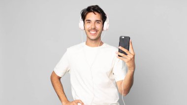 Genç İspanyol adam mutlu bir şekilde gülümsüyor elleri kalçasında ve kulaklıkları ve akıllı telefonlarıyla kendinden emin.