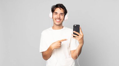 Genç İspanyol adam neşeyle gülümsüyor, mutlu hissediyor ve kulaklıkları ve akıllı telefonlarıyla kenarı işaret ediyor.