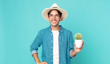 Genç İspanyol adam mutlu bir şekilde gülümsüyor, eli kalçasında, kendinden emin ve elinde bir kaktüs tutuyor.