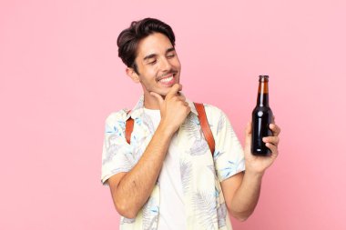 Genç İspanyol bir adam mutlu, kendinden emin bir ifadeyle gülümsüyor. Elleri çenesinde ve elinde bir şişe bira tutuyor.
