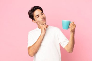 Genç İspanyol adam mutlu bir şekilde gülümsüyor ve hayal kuruyor ya da şüphe ediyor ve elinde kahve fincanı tutuyor.