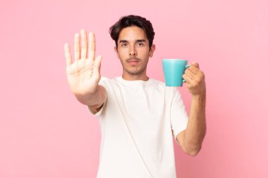Genç İspanyol adam ciddi görünüyor. Avuç içini gösteriyor. El hareketi yapıyor ve elinde kahve fincanı tutuyor.