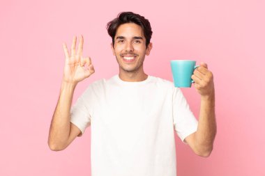 Latin asıllı genç adam mutlu, onaylıyor. İyi bir jest yapıyor ve elinde kahve fincanı tutuyor.