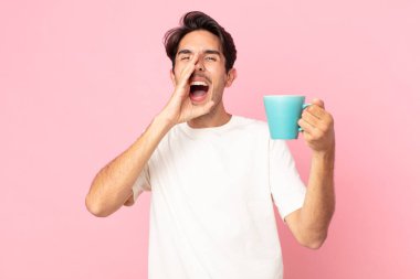 Genç İspanyol adam mutlu hissediyor, elleri ağzının yanında ve elinde kahve fincanı tutarak bağırıyor.