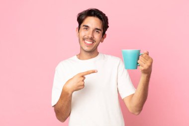 Genç İspanyol adam neşeyle gülümsüyor, mutlu hissediyor ve bir tarafı işaret ediyor ve elinde kahve fincanı tutuyor.
