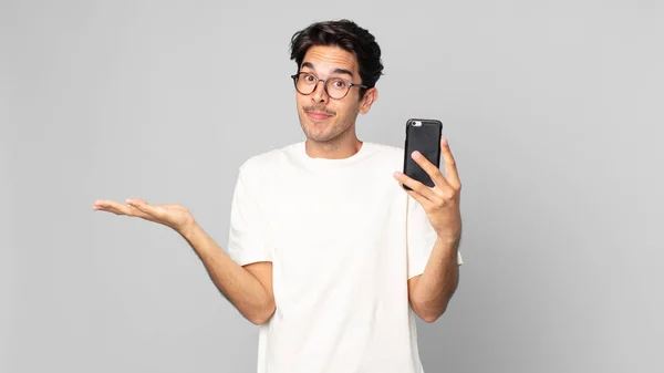 Joven Hispano Sintiéndose Desconcertado Confundido Dudando Sosteniendo Teléfono Inteligente —  Fotos de Stock