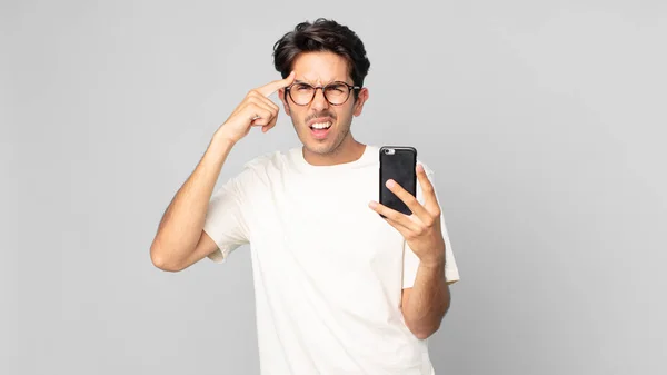 若いヒスパニック系の男は混乱して困惑していて頭がおかしいことを示しスマートフォンを持っています — ストック写真