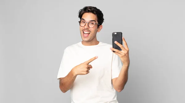 Joven Hispano Mirando Emocionado Sorprendido Señalando Lado Sosteniendo Teléfono Inteligente —  Fotos de Stock