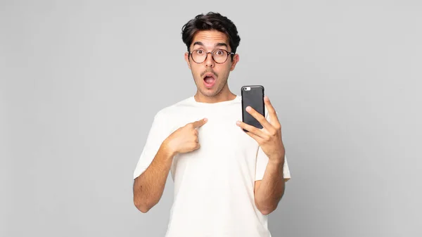 ショックを受けた若いヒスパニック系の男性が口を大きく開けて自分を指差してスマートフォンを持っていて — ストック写真