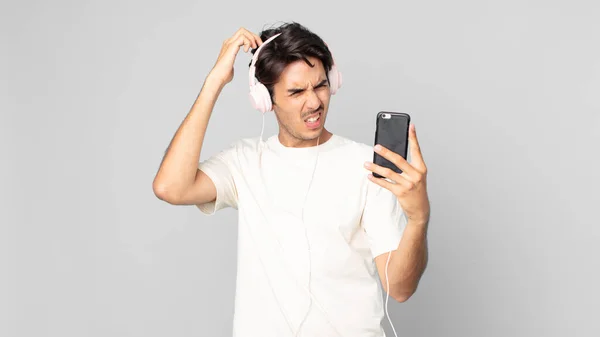 Młody Hiszpan Czuje Się Zakłopotany Zdezorientowany Drapanie Głową Słuchawkami Smartfonem — Zdjęcie stockowe