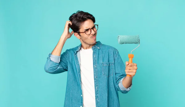 young hispanic man feeling puzzled and confused, scratching head and holding a paint roller