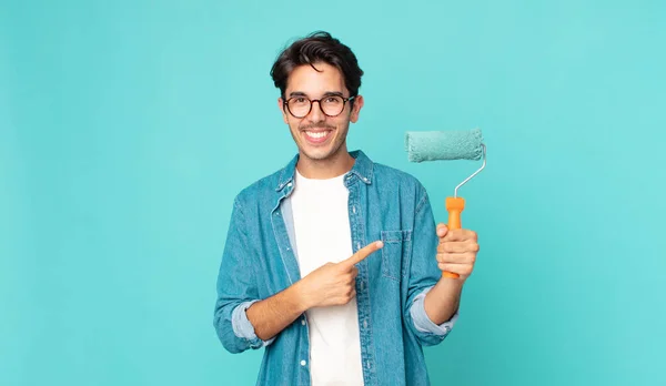 Joven Hispano Sonriendo Alegremente Sintiéndose Feliz Señalando Lado Sosteniendo Rodillo —  Fotos de Stock