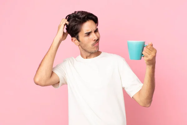 若いヒスパニック系の男は困惑して混乱し頭を掻いてコーヒーマグカップを持っています — ストック写真