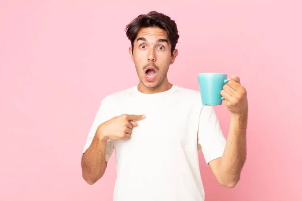 ショックを受けた若いヒスパニック系の男が口を大きく開けて自分を指差してコーヒーマグカップを持っていて — ストック写真