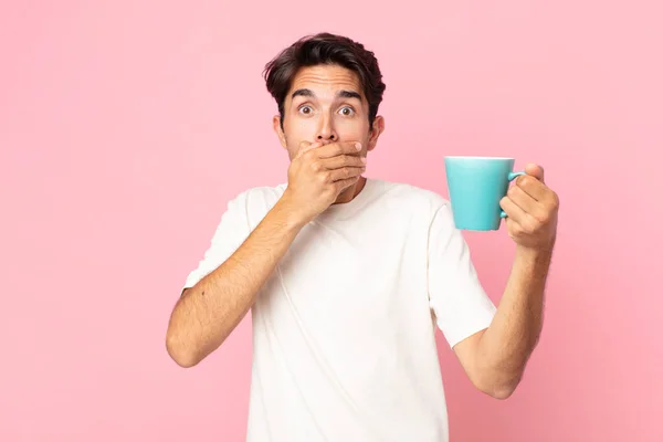 ショックを受けた若いヒスパニック系の男が手で口を覆いコーヒーマグカップを持ち — ストック写真