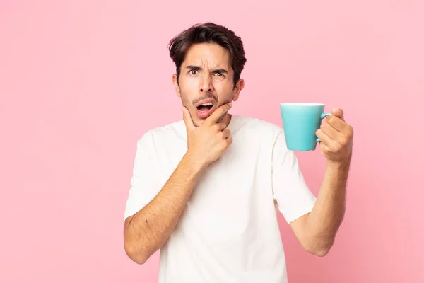 口と目の広い若いヒスパニック系の男が開いて顎を手にし コーヒーマグカップを保持 — ストック写真