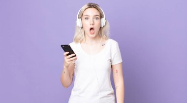Genç, güzel albino kadın kulaklıklar ve akıllı telefonlarla şok olmuş ya da şaşırmış görünüyor.