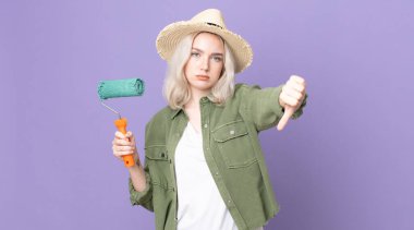 Genç, güzel, albino bir kadın kendini kötü hissediyor, baş parmaklarını gösteriyor ve tekerlekli bir boya tutuyor.