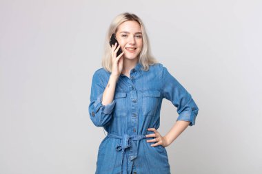 Genç, güzel, albino bir kadın mutlu bir şekilde gülümsüyor, eli kalçasında, kendinden emin ve akıllı bir telefonla konuşuyor.