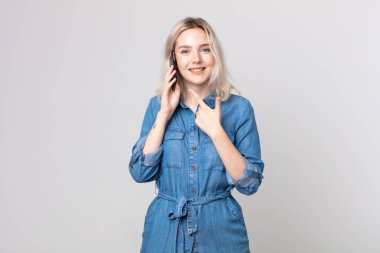 Genç, güzel, albino bir kadın neşeyle gülümsüyor, mutlu hissediyor ve bir tarafı işaret ediyor ve akıllı bir telefonla konuşuyor.