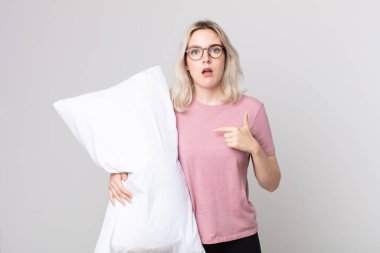 Genç, güzel, albino bir kadın ağzı açık, şok olmuş ve şaşırmış görünüyor. Pijama giymiş ve elinde yastıkla kendini gösteriyor.