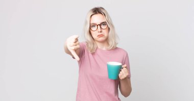 Genç, güzel, albino bir kadın kendini kötü hissediyor. Kahve fincanıyla baş parmağını kaldırıyor.