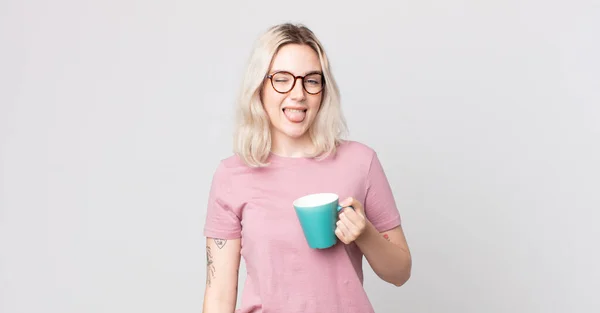 陽気で反抗的な態度の若い可愛いアルビノの女性がコーヒーマグカップで冗談を言いながら舌を出し — ストック写真