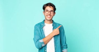 Hispanik yakışıklı adam neşeyle gülümsüyor, mutlu hissediyor ve kenarı ve yukarıyı işaret ediyor, fotokopi uzayında nesne gösteriyor