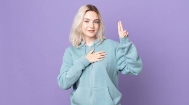 Güzel albino kadın mutlu, kendinden emin ve güvenilir görünüyor, gülümsüyor ve zafer işareti gösteriyor, olumlu bir tavırla.