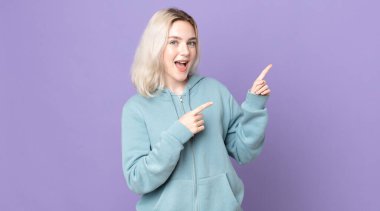 Güzel albino kadın neşeli ve şaşırmış hissediyor, şok olmuş bir ifadeyle gülümsüyor ve bir tarafı işaret ediyor.